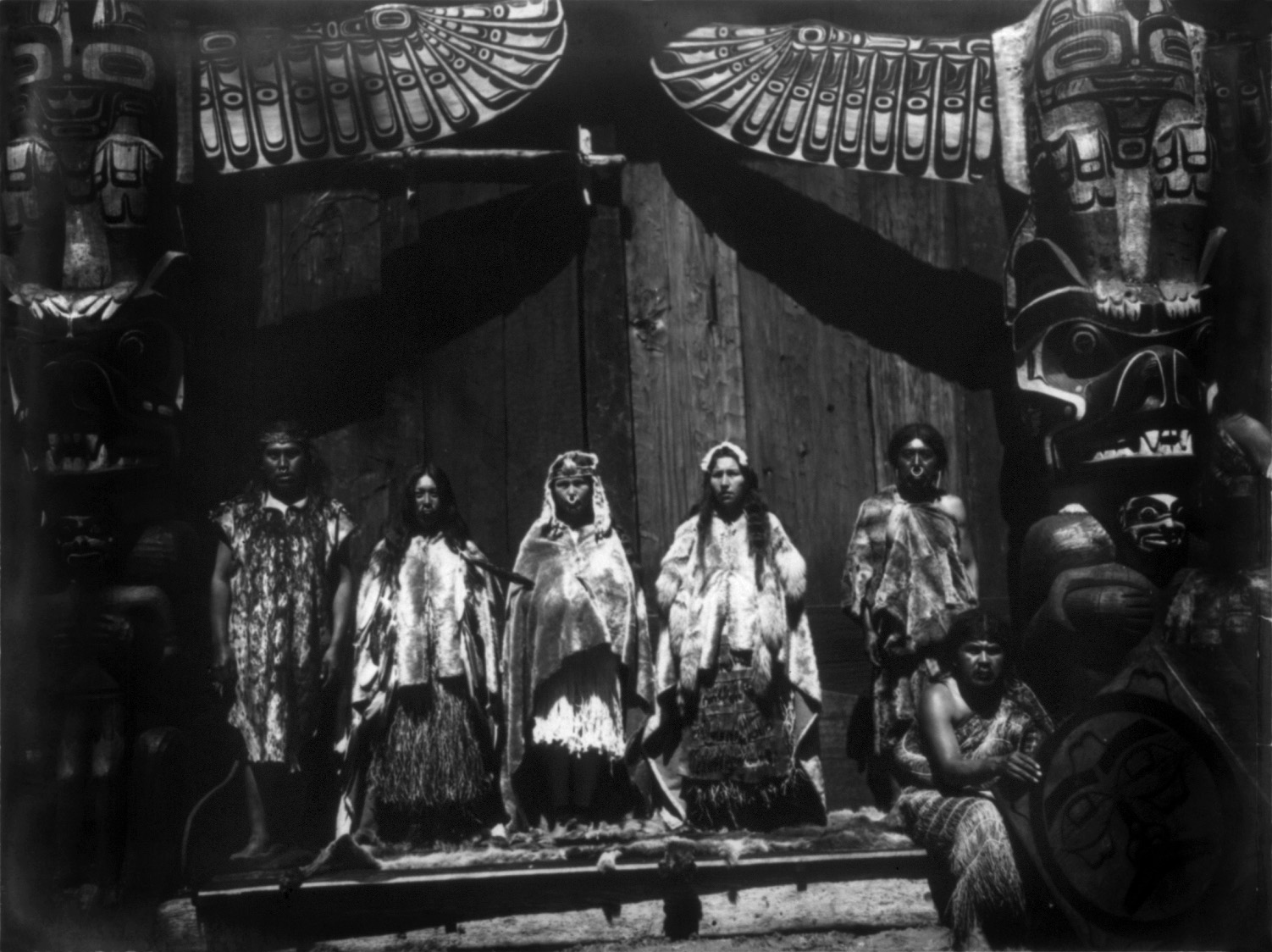 Grupo de personas con mantos de piel de pie sobre una plataforma ante una fachada de tablones pintada con un gran motivo de ave, flanqueada por dos postes tallados