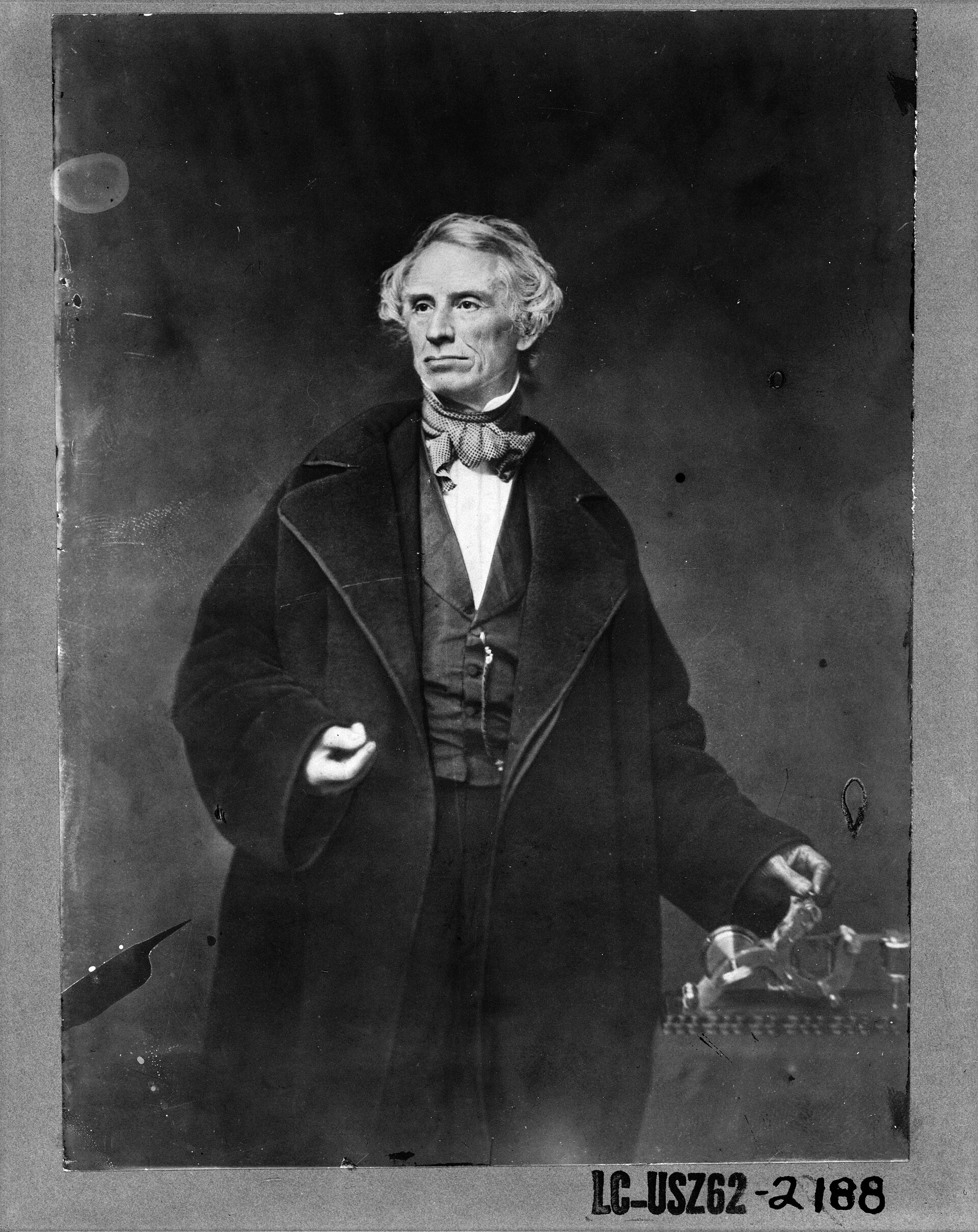 Retrato fotográfico de Samuel Morse, de medio cuerpo, con la mano izquierda apoyada sobre un aparato telegráfico