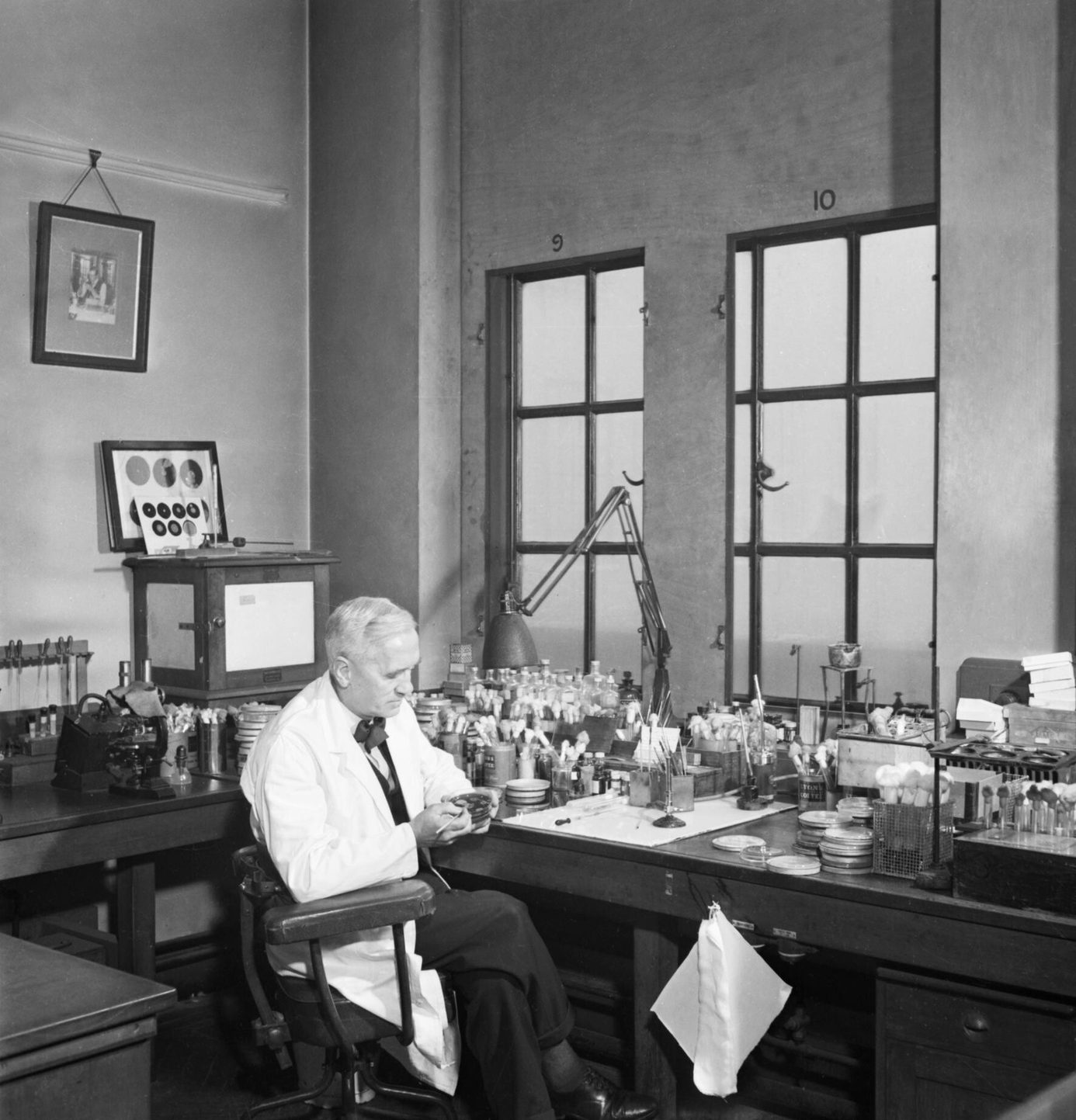 Fotografía en blanco y negro de un hombre mayor con bata blanca sentado a una mesa de laboratorio llena de tubos y frascos, junto a una ventana grande