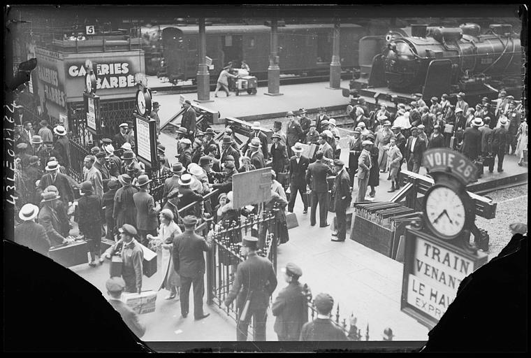 Andén de la estación de Saint-Lazare hacia 1930, con viajeros con sombrero, un gran reloj sobre el cartel de la vía 26 y una locomotora de vapor al fondo; la placa de cristal tiene las esquinas rotas