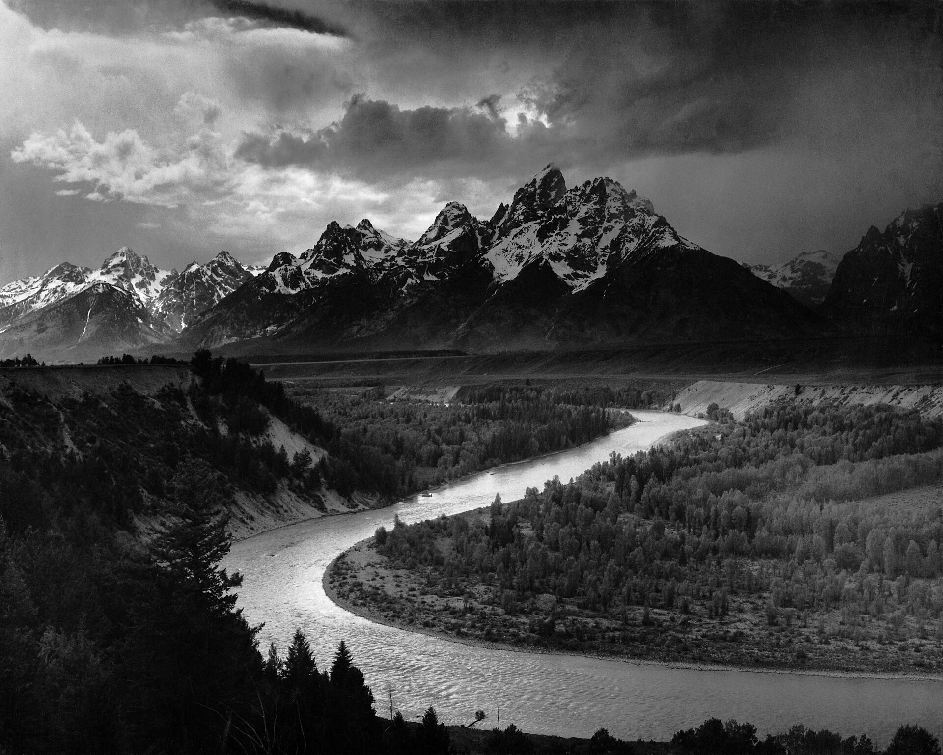 Fotografía en blanco y negro de un río que traza una gran curva brillante a través de un valle boscoso oscuro, con una cordillera nevada y un cielo de nubes al fondo