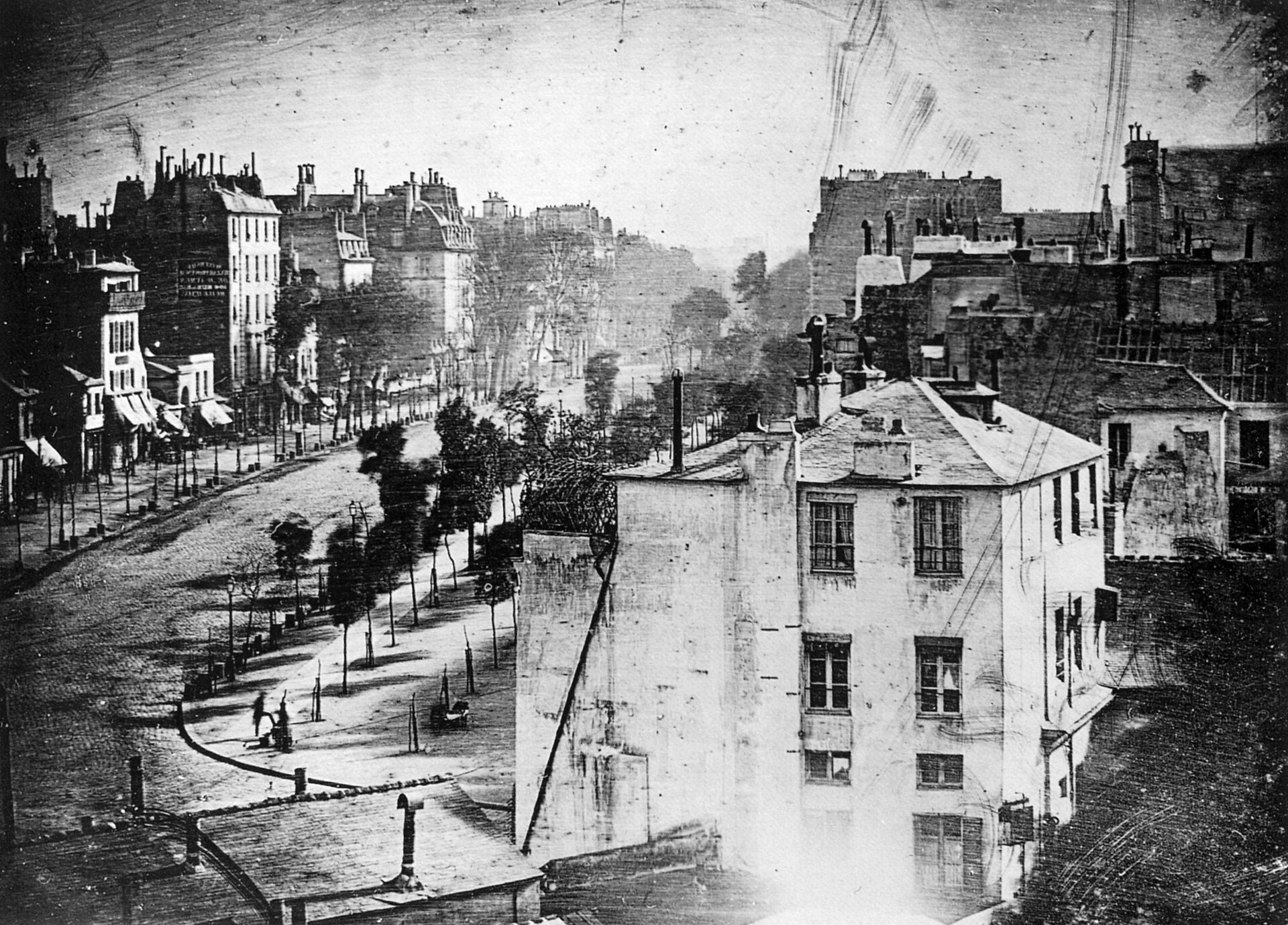 Fotografía antigua en tonos grises de una avenida arbolada vista desde lo alto, con edificios de varias plantas a la izquierda y una casa en primer plano; la calzada parece vacía