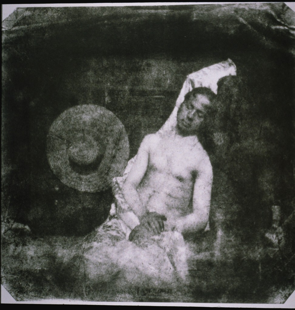 Fotografía antigua en tonos sepia de un hombre con el torso desnudo, sentado y recostado de lado contra una pared, con los ojos cerrados y una tela clara sobre las piernas