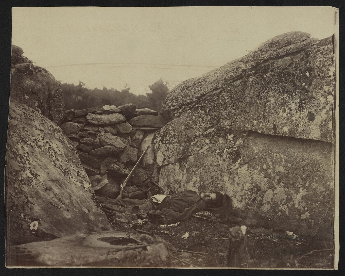 Fotografía de 1863 de un soldado muerto tendido boca arriba entre dos grandes rocas, con un murete de piedras detrás y un fusil apoyado en la pared