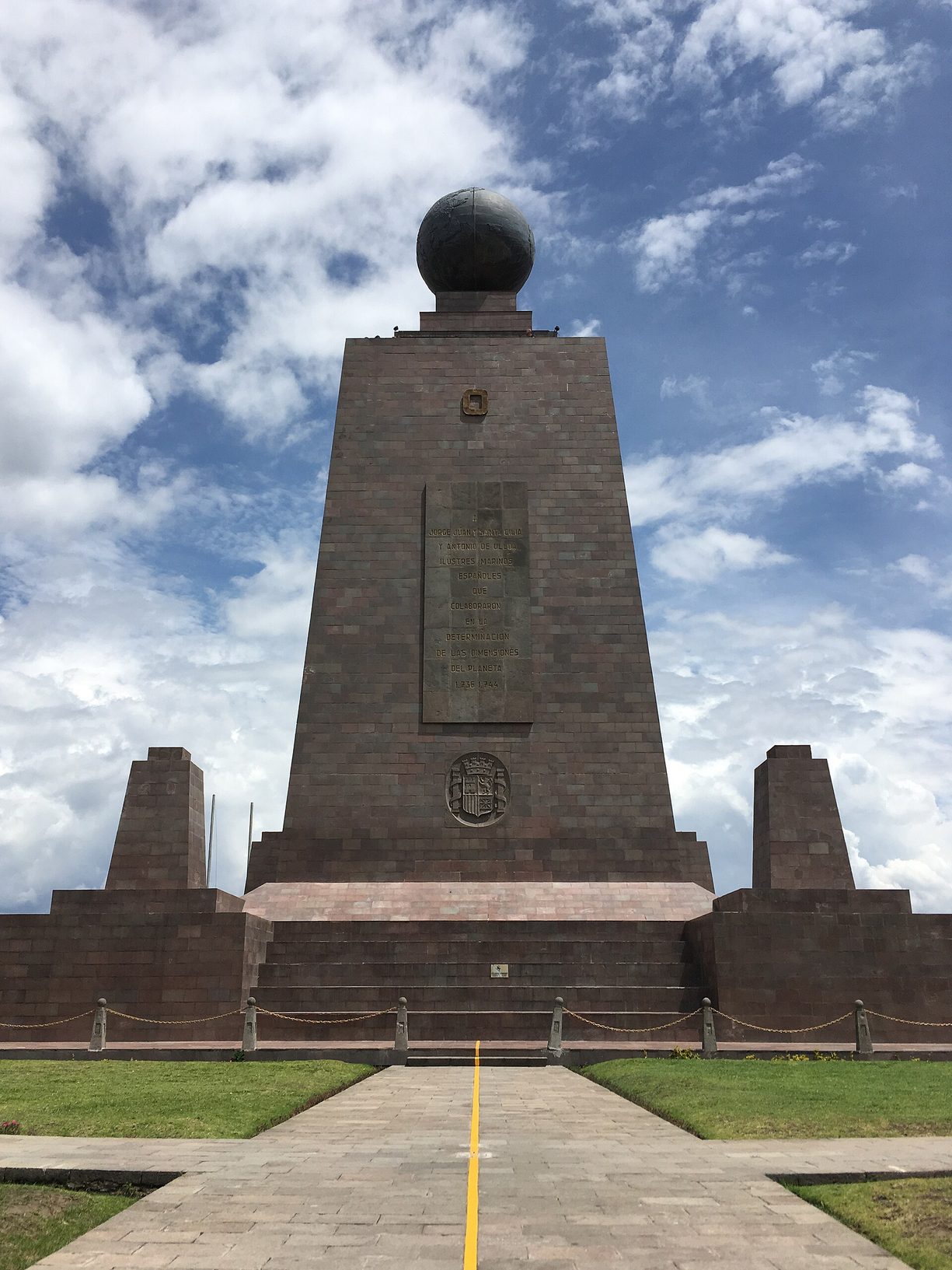 Monumento de piedra en forma de pirámide truncada coronado por un globo terráqueo, con una franja amarilla en el suelo que conduce hasta él