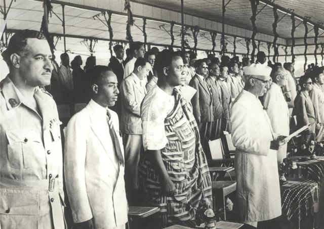 Fotografía en blanco y negro de una fila de delegados de pie bajo un toldo; a la izquierda un hombre con uniforme militar y bigote, en el centro uno vestido con una tela africana estampada y a la derecha un hombre mayor con gorro blanco