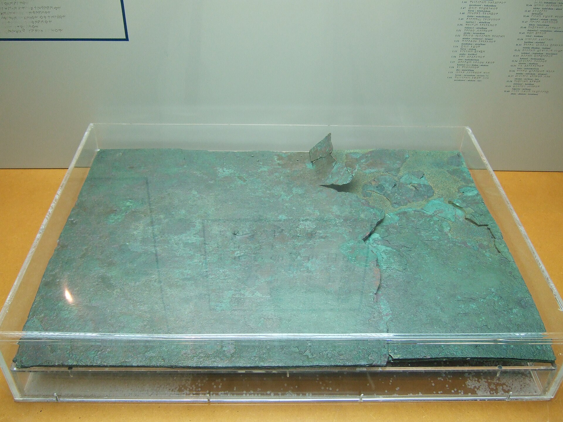Gran placa de bronce verdosa y fragmentada, expuesta en horizontal dentro de una vitrina de metacrilato