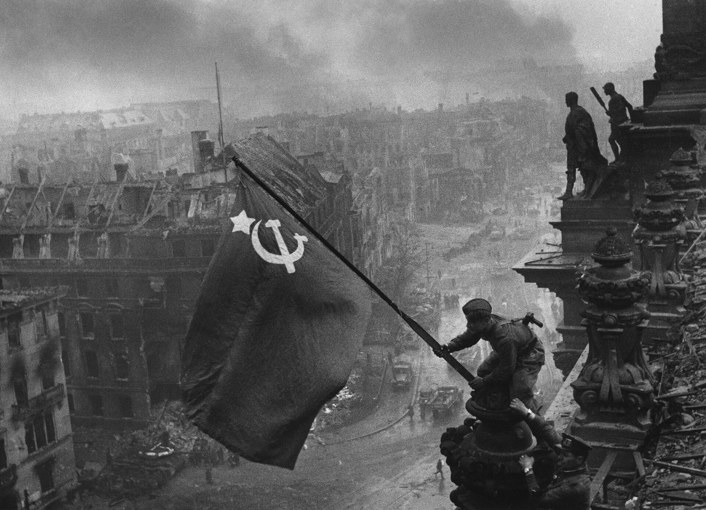 Fotografía en blanco y negro de un soldado que coloca una bandera con la hoz y el martillo en lo alto del Reichstag, sobre una ciudad en ruinas cubierta de humo