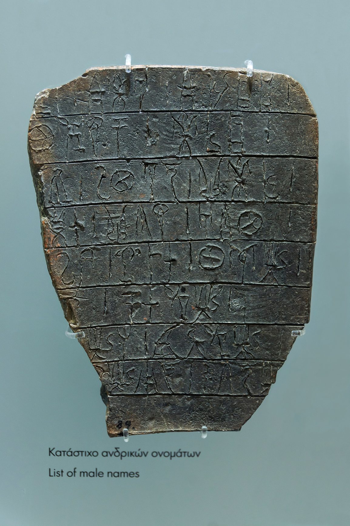 Tablilla de arcilla oscura de forma rectangular irregular, cubierta de varias líneas de signos incisos, expuesta en una vitrina de museo con una cartela bilingüe debajo