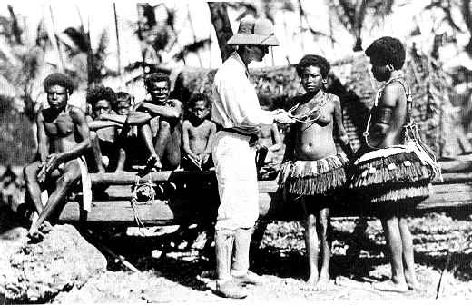 Fotografía en blanco y negro de un hombre con sombrero y ropa clara que conversa con varias personas trobriandesas, algunas con faldas de fibra vegetal, junto a unas casas