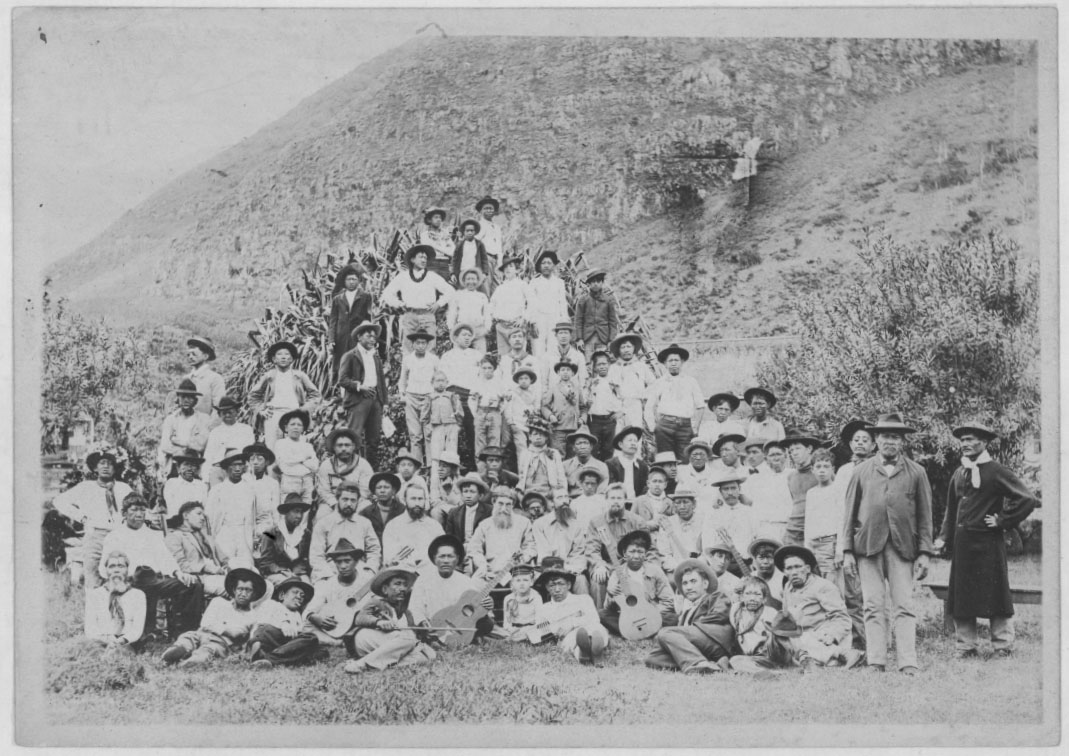 Fotografía antigua en blanco y negro de un grupo numeroso de hombres, mujeres y niños posando al pie de una ladera empinada