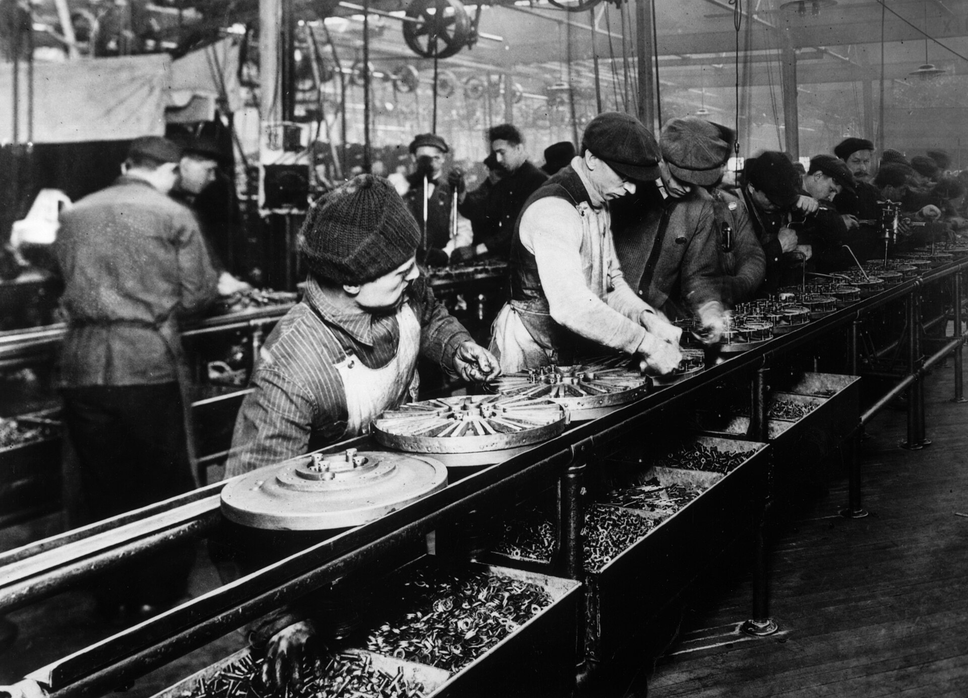 Fotografía en blanco y negro de una larga fila de obreros con gorra trabajando a ambos lados de una cinta de montaje sobre la que avanzan piezas mecánicas